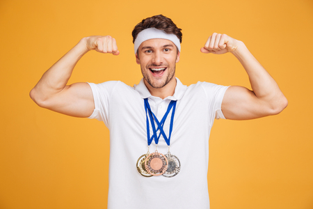 Cheerful young spotrsman with three medals showing biceps over yellow backgroundの写真素材