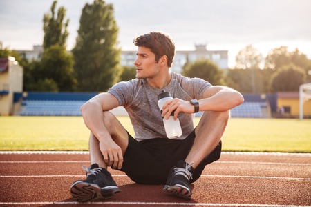 Young handsome man athlete resting at the stadium and drinking water, looking awayの写真素材