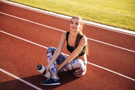 Fitness young woman sitting on running track and holding water bottle at the stadiumの写真素材