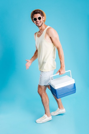 Smiling young man in hat and sunglasses walking and holding cooler bagの写真素材