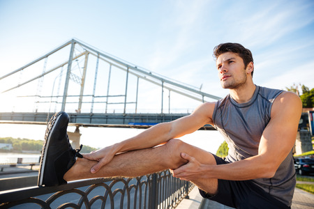 Handsome young sports man doing stretching leaning against bridge railing and looking away outdoors
