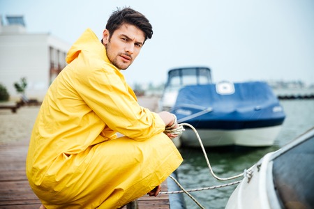 Handsome young sailor in yellow cloak holding rope at the pier and looking at cameraの写真素材