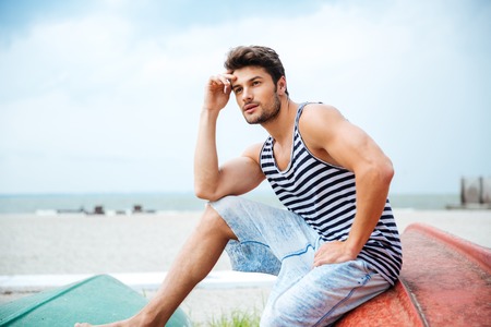 Pensive young man sitting on a fishing boat and looking away by the seaの写真素材