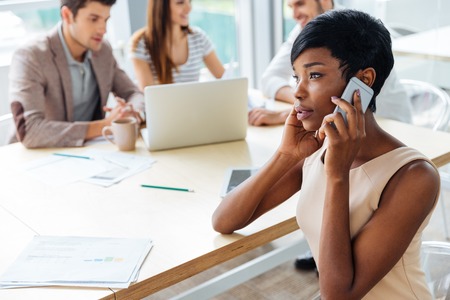 Successful african american young businesswoman sitting at the table and talking on mobile phone in officeの写真素材
