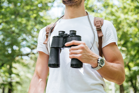 Closeup of young man with backpack standing and holding binoculars in forestの写真素材
