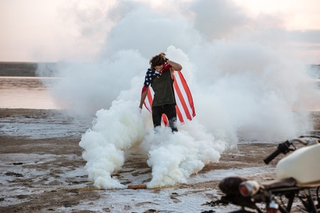 Brutal man wearing usa flag cape posing in white smoke outdoors