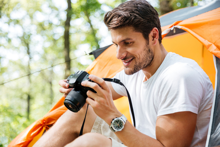 Cheerful young man tourist sitting and taking photos with modern photo camera in forestの写真素材
