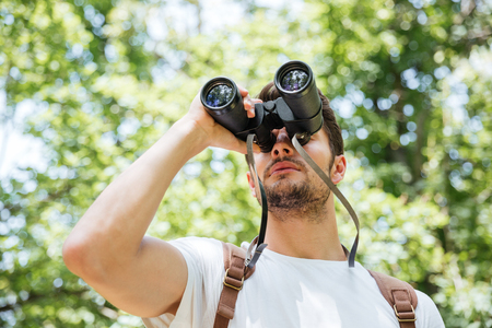 Concentrated young man with backpack looking through binoculars in forestの写真素材