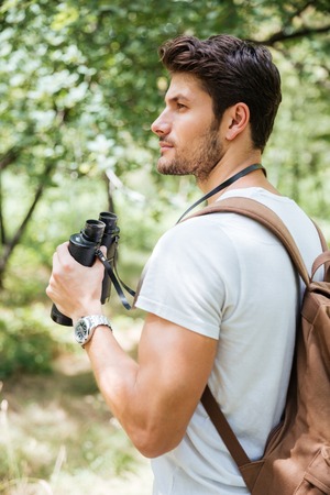 Pensive young man with backpack holding binoculars and thinking in forest