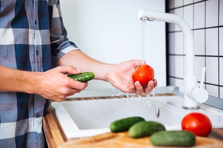 Closeup of young man in checkered shirt washing tomatoes and cucumbers on the kitchenの写真素材