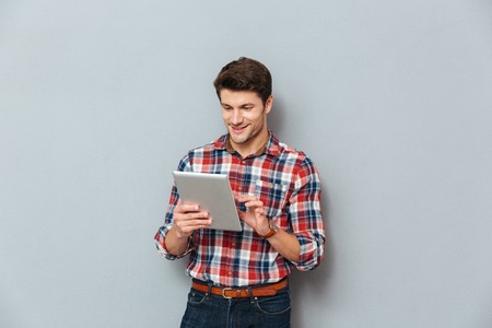 Happy young man in plaid shirt standing and using tablet over grey background