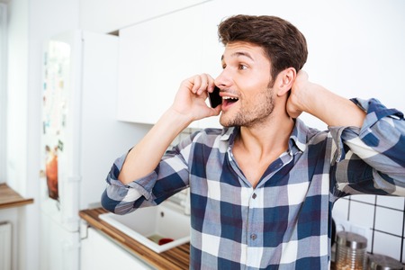 Happy amazed young man in checkered shirt talking on cell phone on the kitchenの写真素材