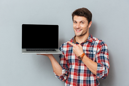 Happy young man in plaid shirt pointing on blank screen laptop over grey backgroundの写真素材