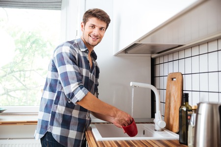 Happy young man in plaid shirt standing and washing dishes on the kitchenの写真素材