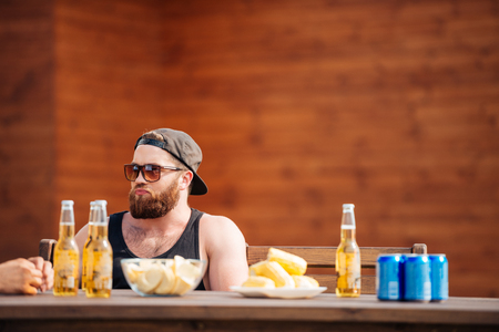 Handsome bearded young man in cap and sunglasses drinking beer at the table outdoorsの写真素材