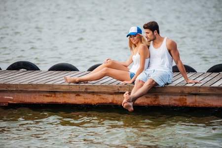 Beautiful young couple sitting barefoot and talking on pierの写真素材