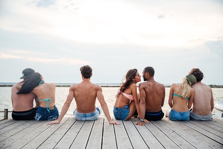 Back view of happy young couples sitting and hugging on pier