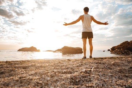 Young handsome man athlete standing with arms raised at the rocky beach by the sea looking at sunset