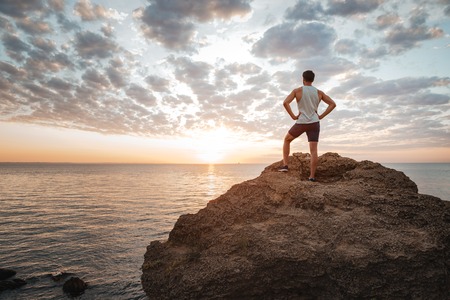 Young sportsman standing backwards on the mountain rock by the sea and looking at sunsetの写真素材