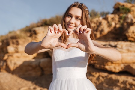 Young smiling bride showing heart gesture with hands standing at the beachの写真素材