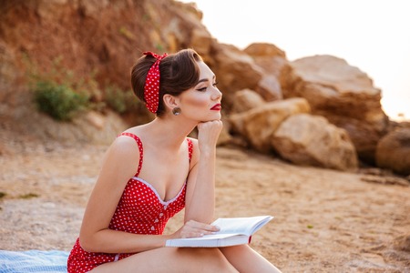 Young beautiful pensive pin up girl sitting with book at the beachの写真素材