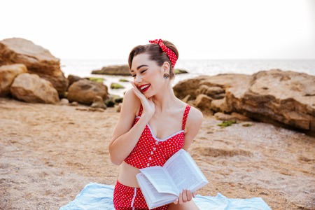 Young cute pin up woman with bright make up looking away and holding book while sitting on the beachの写真素材