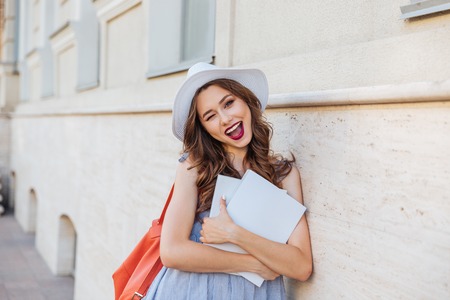 Cheerful pretty young woman in hat with blank covered magazines winking and showing thumbs upの写真素材
