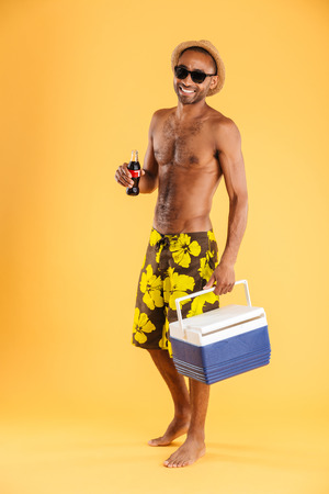 Cheerful young man in hat and sunglasses holding cooler bag and drinking coke over orange backgroundの写真素材