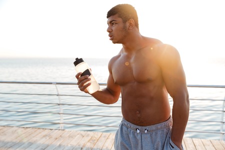Muscular african american young man athlete standing on pier and holding bottle of waterの写真素材