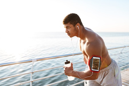 Concentrated african american young man athlete with blank screen smartphone in armband working out and drinking waterの写真素材