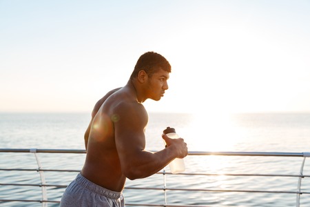 Focused shirtless african american young sportsman with bottle of water running on pier