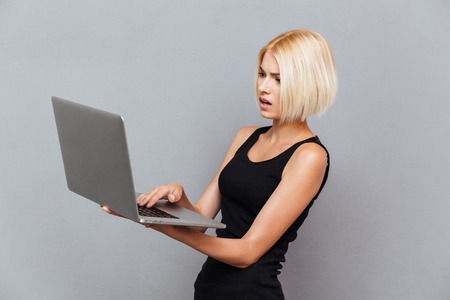 Concentrated beautiful young woman hoding and using laptop over gray backgroundの写真素材