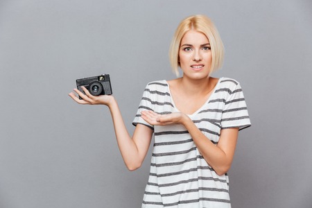 Confused blonde young woman holding old vintage photo camera over gray background