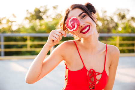 Cheerful playful young woman covered her eye with lollipop standing outdoorsの写真素材