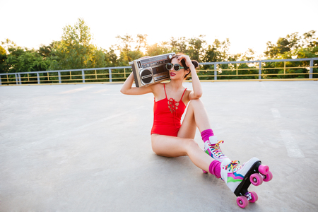 Young pretty brunette roller girl in sunglasses and red swimsuit listening music with old record player while sitting on the open roadの写真素材