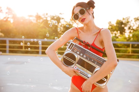 Pretty young woman in sunglasses and red swimsuit standing outdoors and holding old vintage boomboxの写真素材