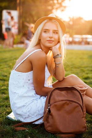 Beautiful blonde young woman in hat with backpack sitting in the parkの写真素材