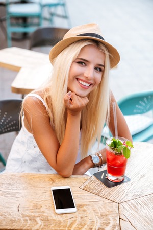 Happy pretty young woman with blank screen mobile phone drinking cocktail in outdoor cafeの写真素材