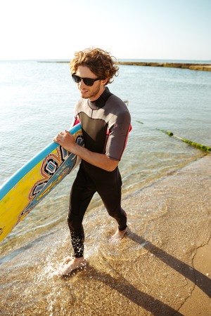 Surfer man walking with surf board in his hands across the sea shore