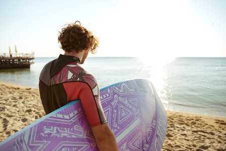 Back view of curly young man surfer with surfing board on the beachの写真素材