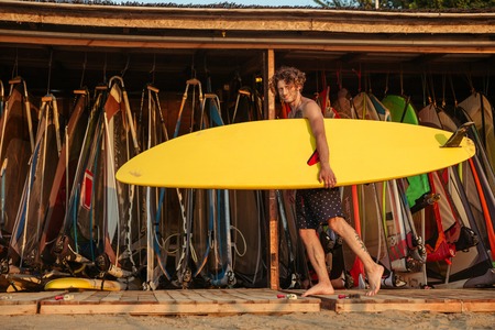 Young handsome curly surfer holding surf board while standing at the shack outdoorsの写真素材