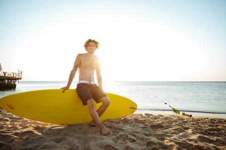 Young handsome curly resting while sitting on the surfboard at the beachの写真素材