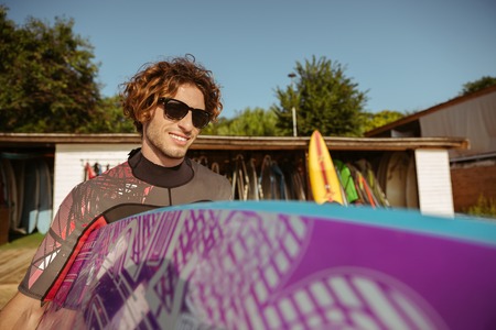 Young handsome curly man in sunglasses going to surf holding surfboard