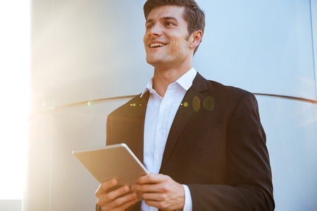 Cheerful attractive young businessman standing outdoors and using pc tabletの写真素材
