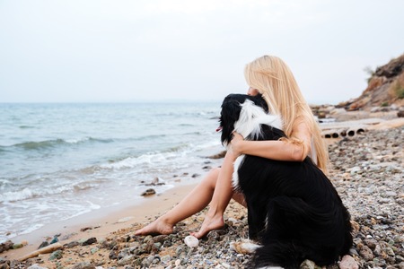 Blonde young woman sitting and hugging a dog on the beachの写真素材