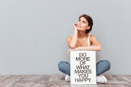 Smiling woman sitting on the floor and holding board with motivational phrase isolated on gray backgroundの写真素材