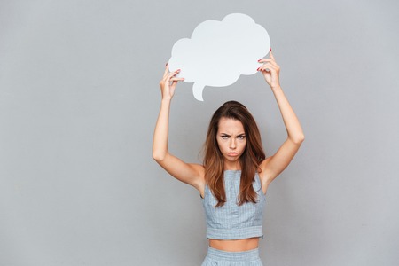 Sad frowning young woman standing and holding empty speech bubble over grey backgroundの写真素材