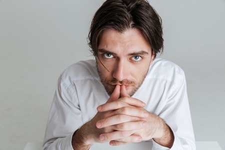 Close up portrait of a young brunette man sitting with fingers crossed isolated on the gray backgroundの写真素材