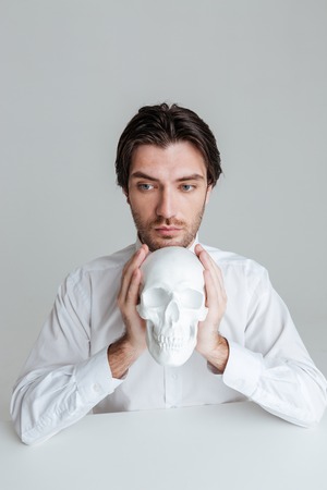 Close up portrait of a man in white shirt holding fake skull at his face isolated on the gray backgroundの写真素材