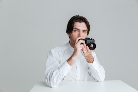 Young bristled man ready to make a photo with camera while sitting at the table isolated on the gray...